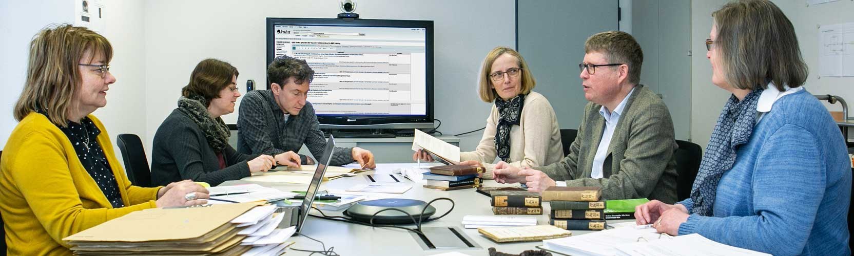 Library team meeting at a round table, in the background a large screen showing the library catalog